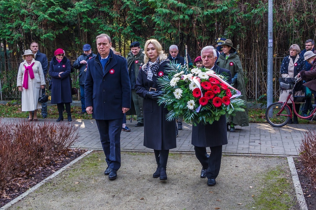 Park. Dwóch mężczyzn i jedna kobieta stoją obok siebie w rzędzie. Jeden z mężczyzn w rękach trzyma duży wieniec z biało-czerwonymi kwiatami.