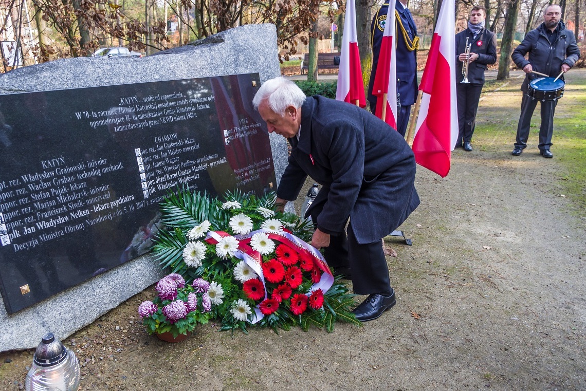 Park. Mężczyzna w średnim wieku składa wieniec z biało-czerwonymi kwiatami przed pomnikiem Pamięci Ofiar Katynia. 
