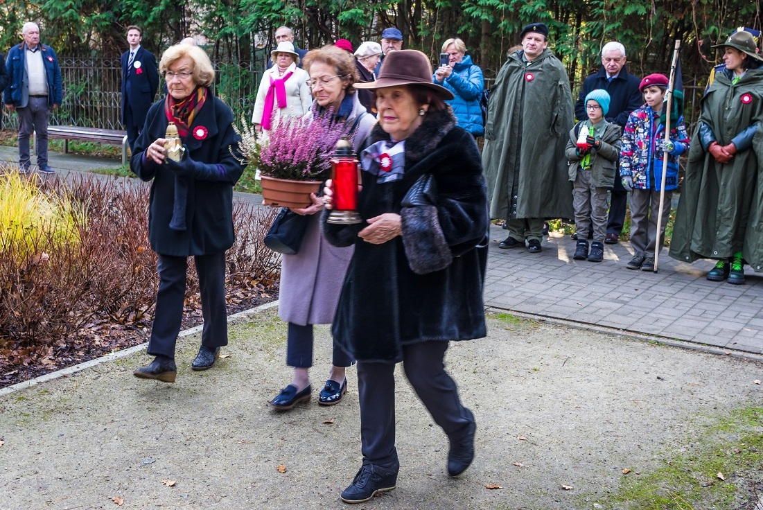 Park. Trzy seniorki idą złożyć kwiaty i znicze przed pomnikiem Pamięci Ofiar Katynia.