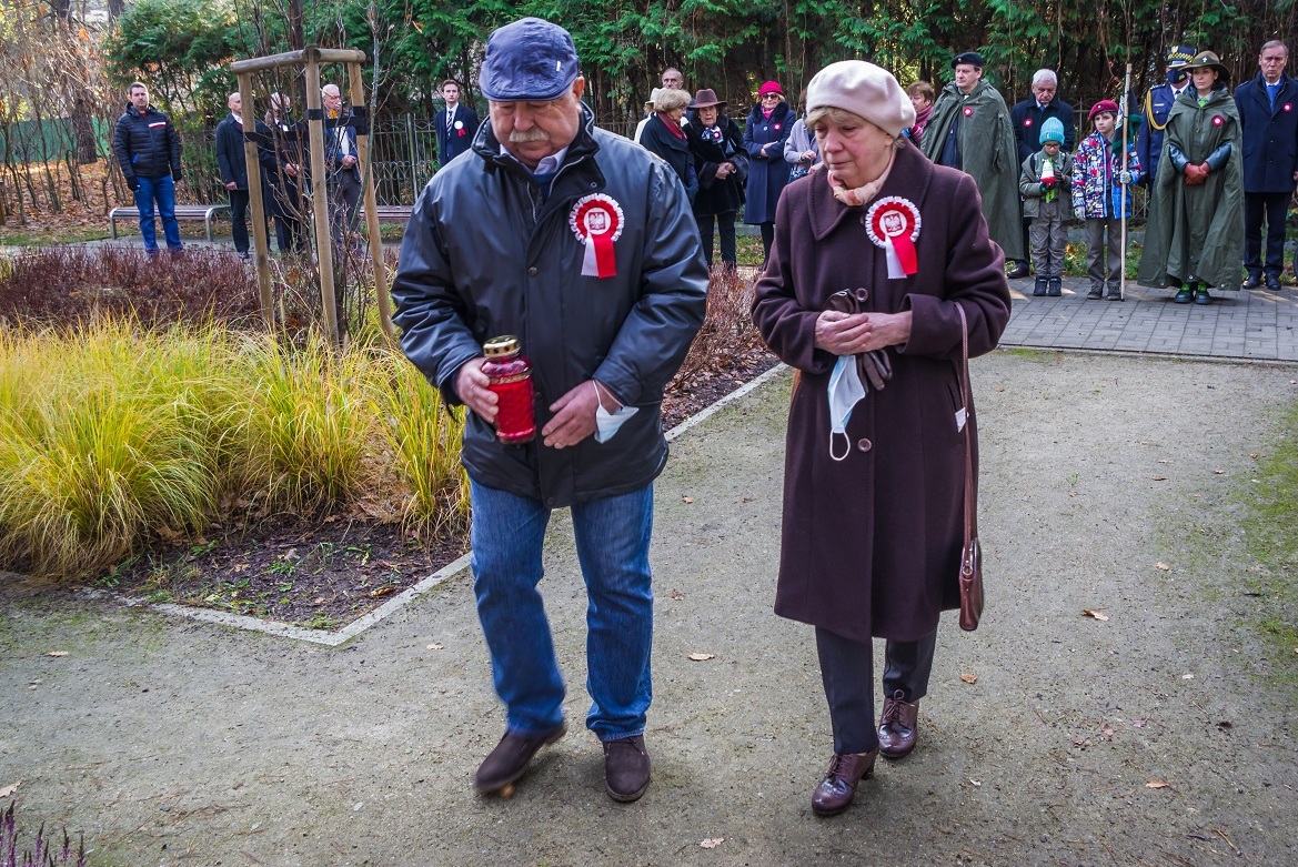 Park. Para seniorów, kobieta i mężczyzna składają znicze przed pomnikiem Pamięci Ofiar Katynia.