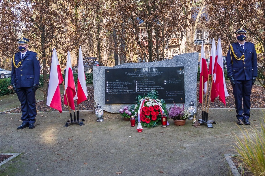 Przy pomniku Pamięci Ofiar Katynia stoi dwóch mężczyzn, którzy ubrani są w mundury Straży Miejskiej. Obok pomnika ustawione są biało-czerwone flagi oraz stoją kwiaty i znicze.