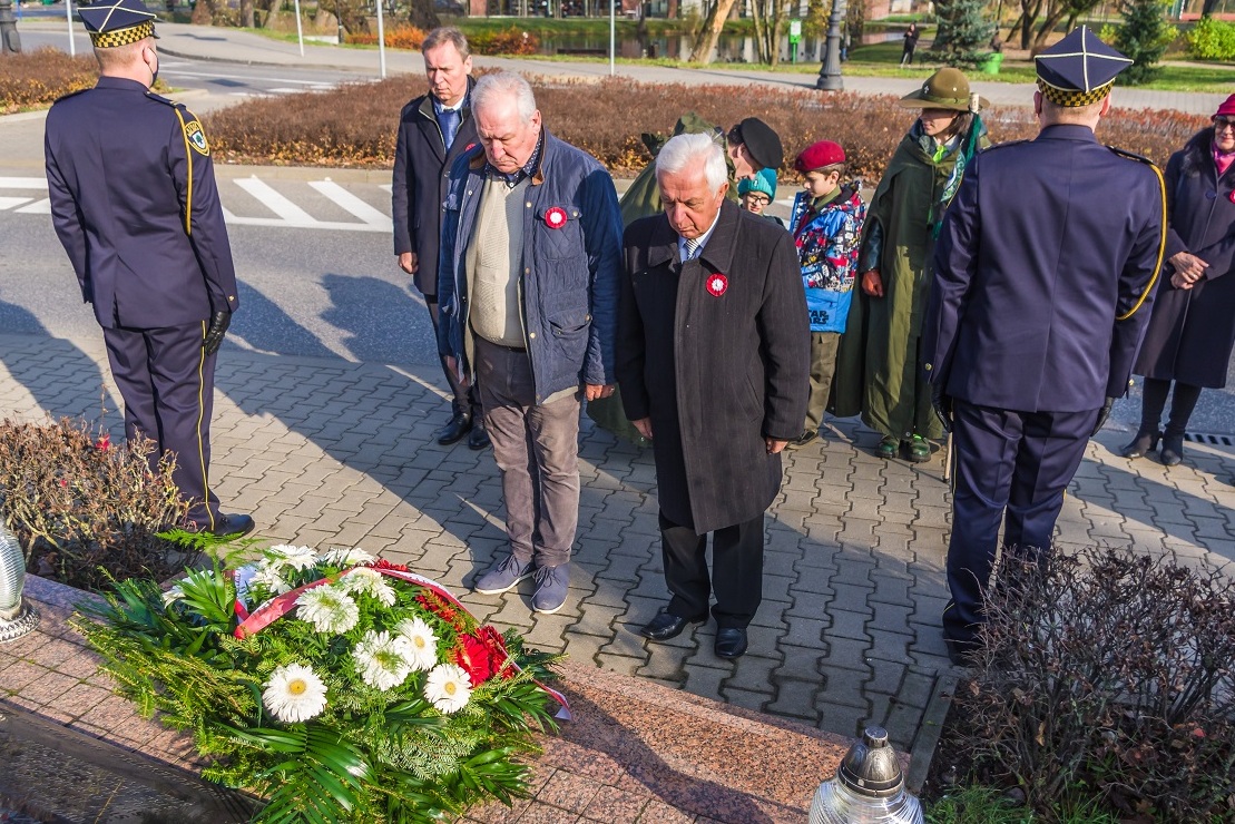 Trzech mężczyzn w średnim wieku stoi przy pomniku. Na pomniku leży wiązanka z biało-czerwonymi kwiatami.