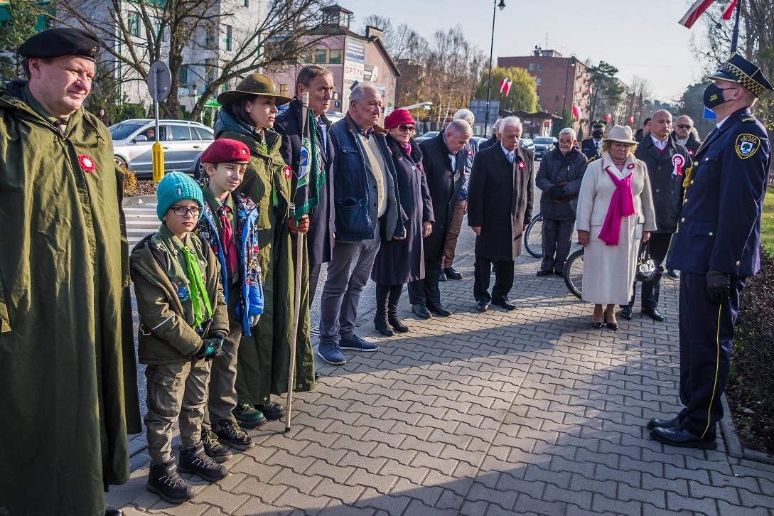 Około piętnastu osób stoi obok siebie w rzędzie na chodniku. W tle widać ulicę i budynki. 