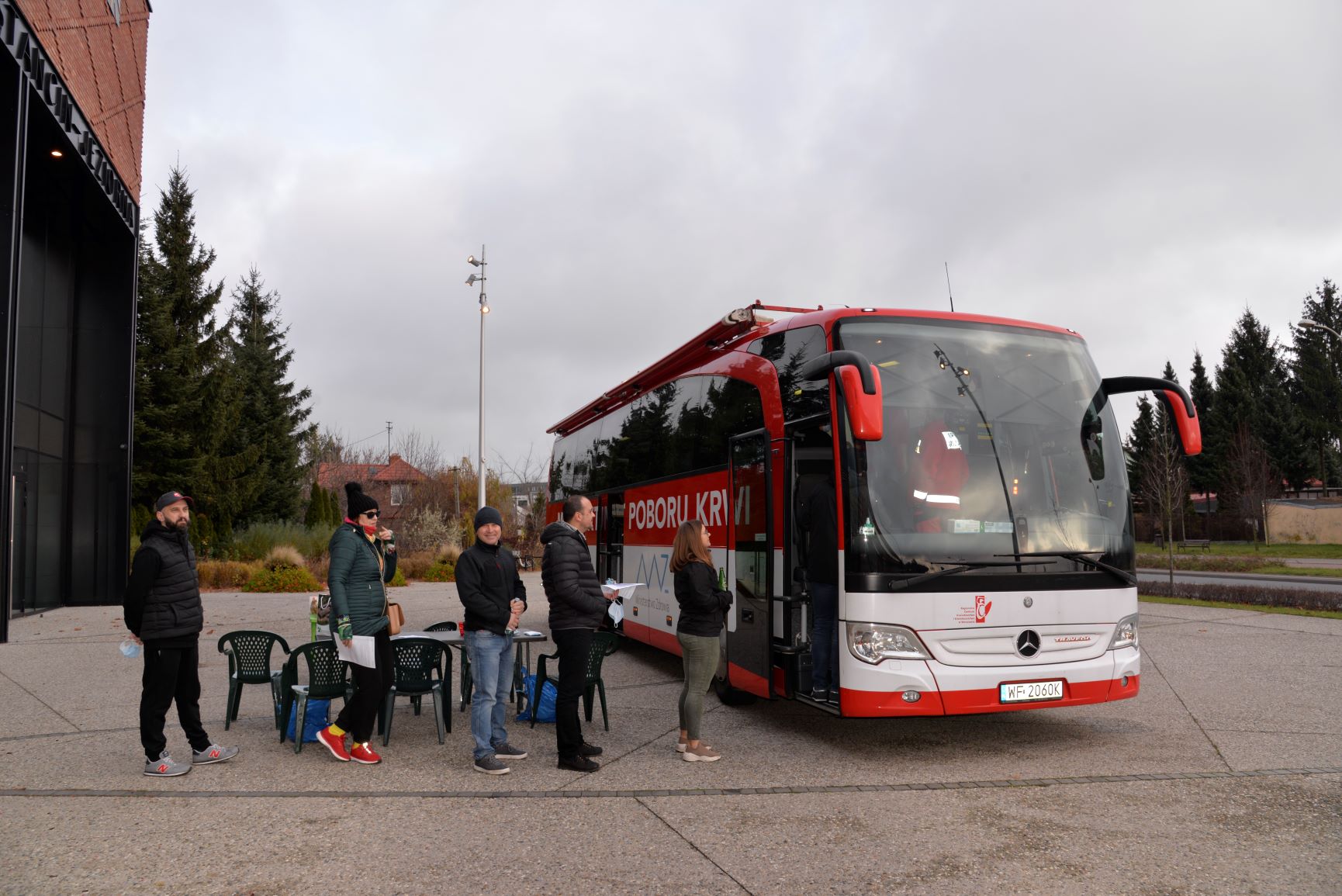 Na chodniku stoi duży czerwony autokar, który ma z boku duży biały napis Mobilny Punkt Poboru Krwi. Przed autokarem w kolejce stoi pięć osób.