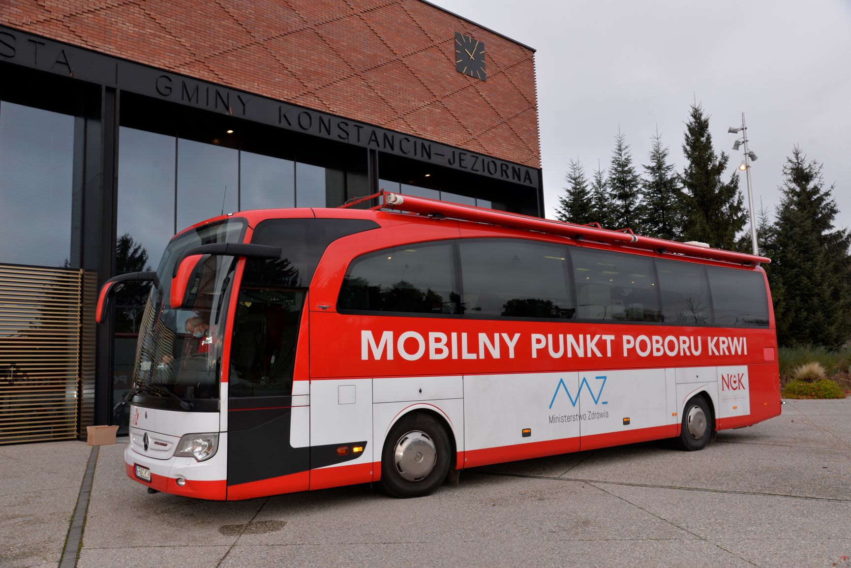 Na chodniku stoi duży czerwony autokar, który ma z boku duży biały napis Mobilny Punkt Poboru Krwi. Niżej jest logo i napis Ministerstwo Zdrowia. W tle jest budynek Urzędu Miasta i Gminy Konstancin-Jeziorna.