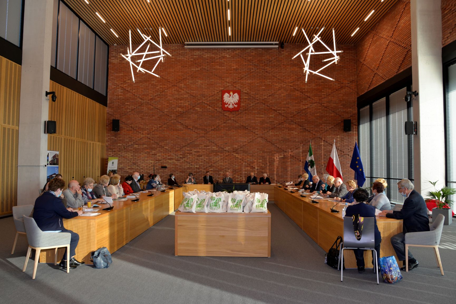 Sala posiedzeń. Przy drewnianych stołach ustawionych w literę U na krzesłach siedzi około trzydzieści osób. W tle jest ściana z czerwonej cegły. 
