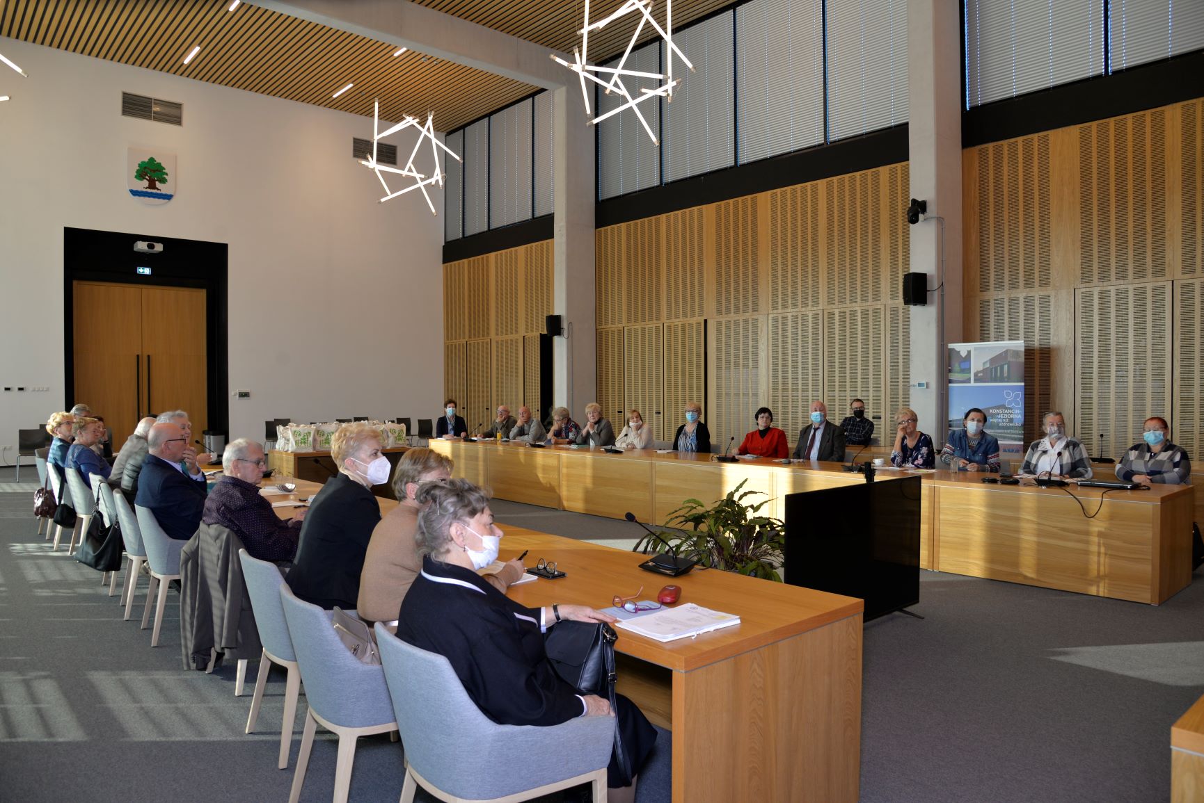 Sala posiedzeń. Przy dwóch drewnianych stołach ustawionych naprzeciwko siebie siedzi około trzydzieści osób, seniorzy i seniorki. 