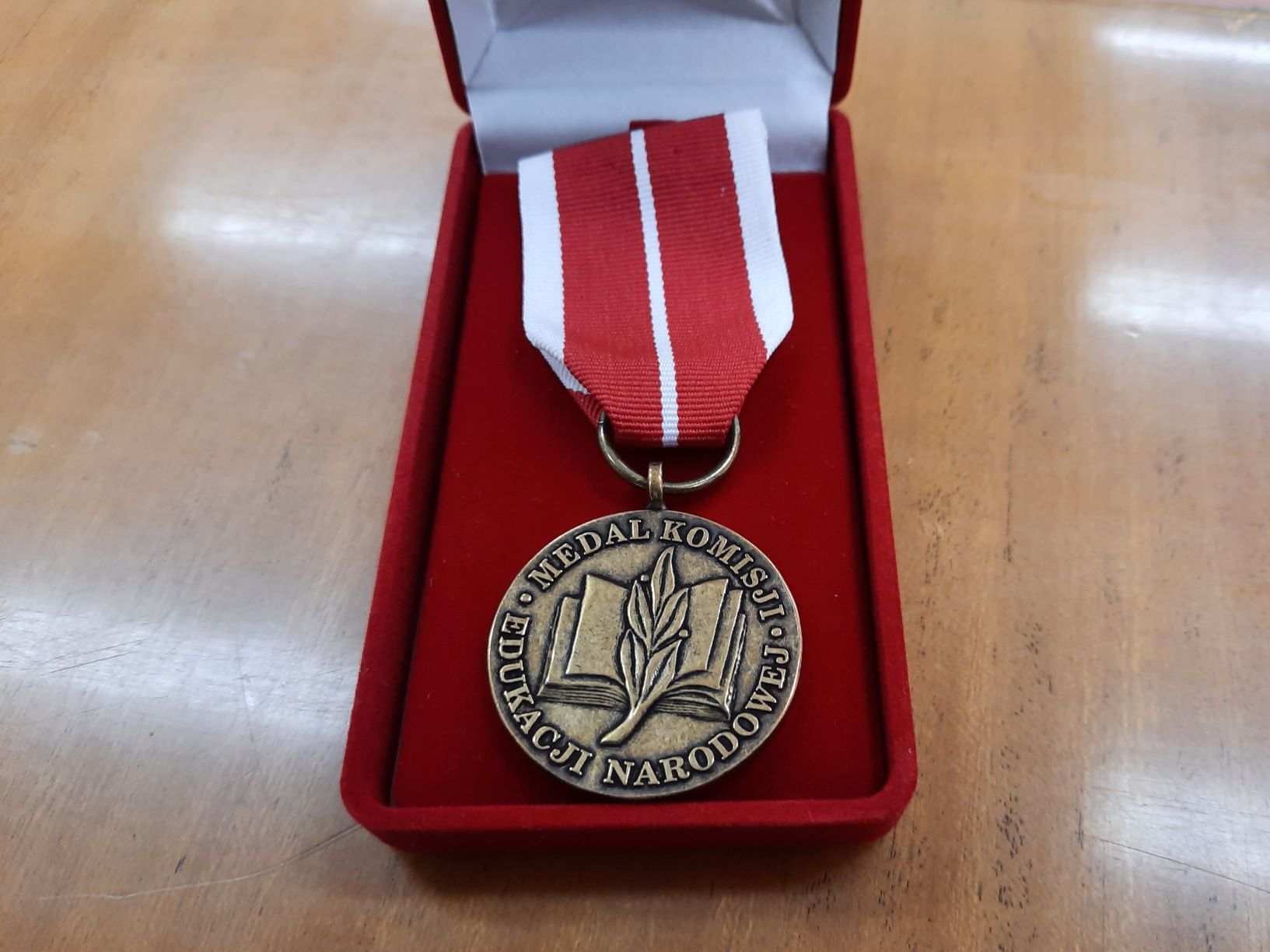 W czerwonym etui leży medal, który ma kształt koła. Wykonany jest z metalu koloru jasnobrązowego. Na nim widnieje wizerunek otwartej książki przełożonej gałązką drzewa laurowego oraz napis w Medal Komisji Edukacji Narodowej.