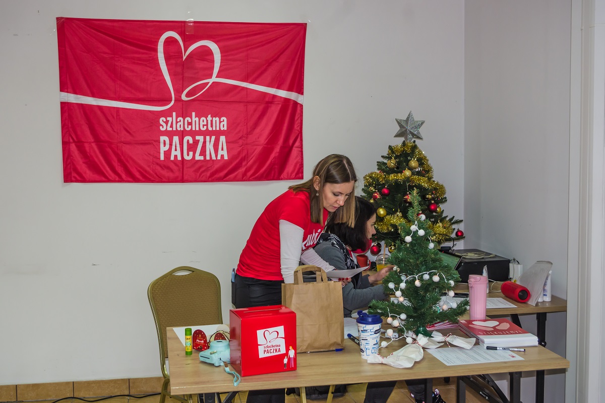 Biurko. Przy nim stoi kobieta, która pracuje na komputerze. Za nią stoi choinka, a na ścianie wisi czerwony baner z napisem: szlachetna paczka. 