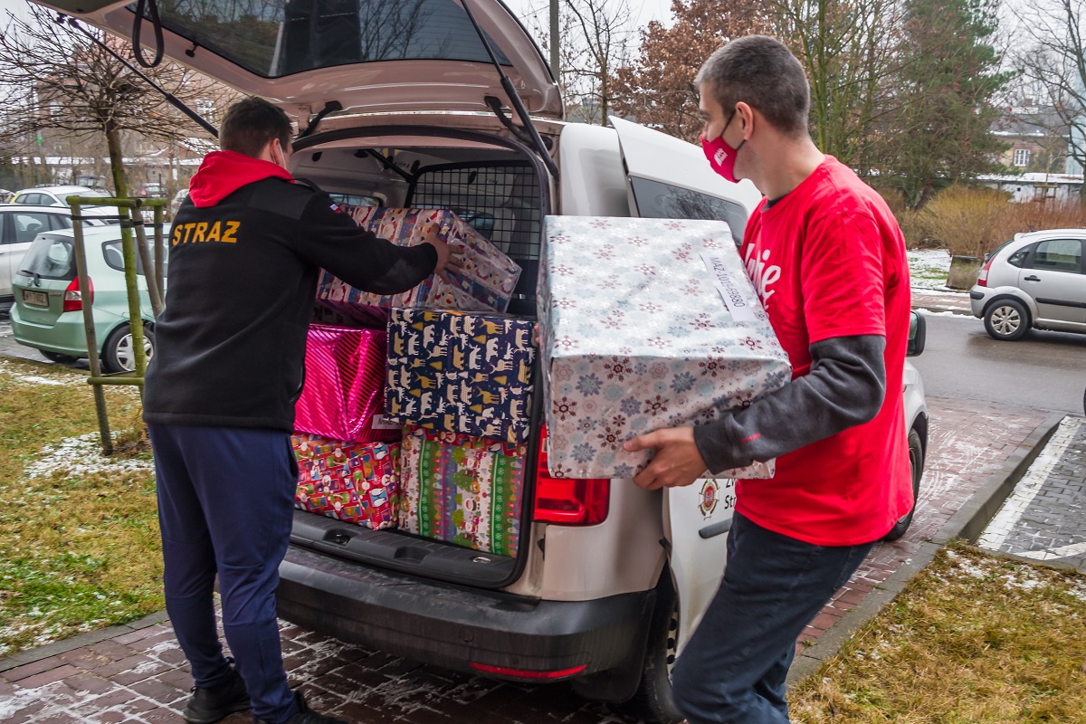 Samochód dostawczy, który ma otwarty bagażnik. Znajdują się w nim duże pudła – prezenty świąteczne. Obok samochodu stoi dwóch mężczyzn, jeden pokuje do bagażnika pudełka, drugi mu je podaje. 