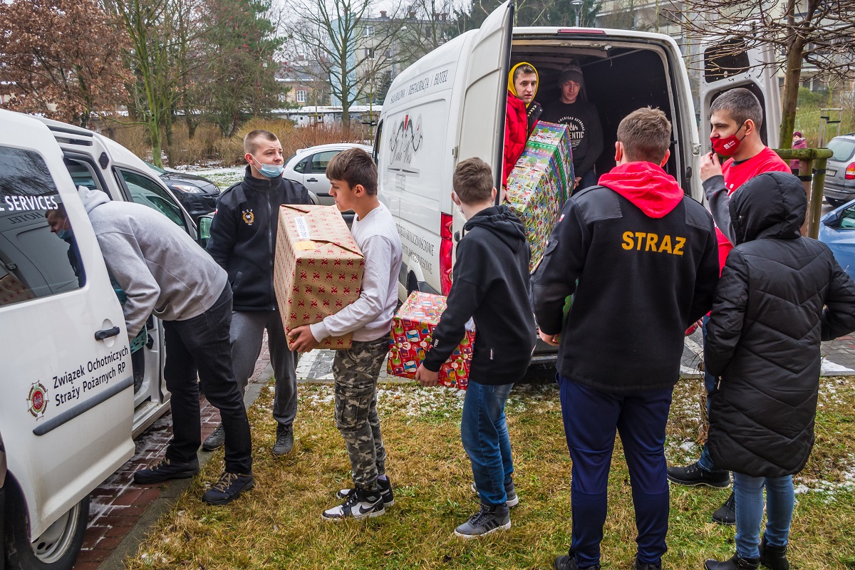 Dwa samochody dostawcze stoją obok siebie. Mają otwarte bagażniki. 9 osób, przepakowuje duże pudła – prezenty świąteczne z jednego samochodu do drugiego. 