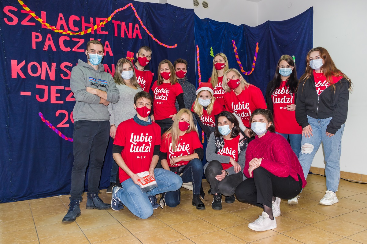 Zdjęcie grupowe – 14 osób stoi obok siebie w trzech rzędach. Większość ma na sobie czerwoną koszulkę z dużym napisem: lubię ludzi.