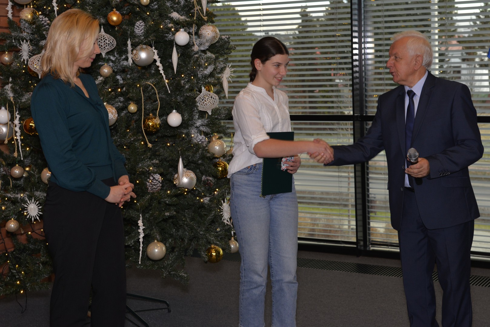 Trzy osoby stoją na tle choinki (dwie kobiety – starsza i młodsza oraz mężczyzna). Mężczyzna (burmistrz) gratuluje stojącej w środku młodej kobiecie, trzymającej w ręku list gratulacyjny.