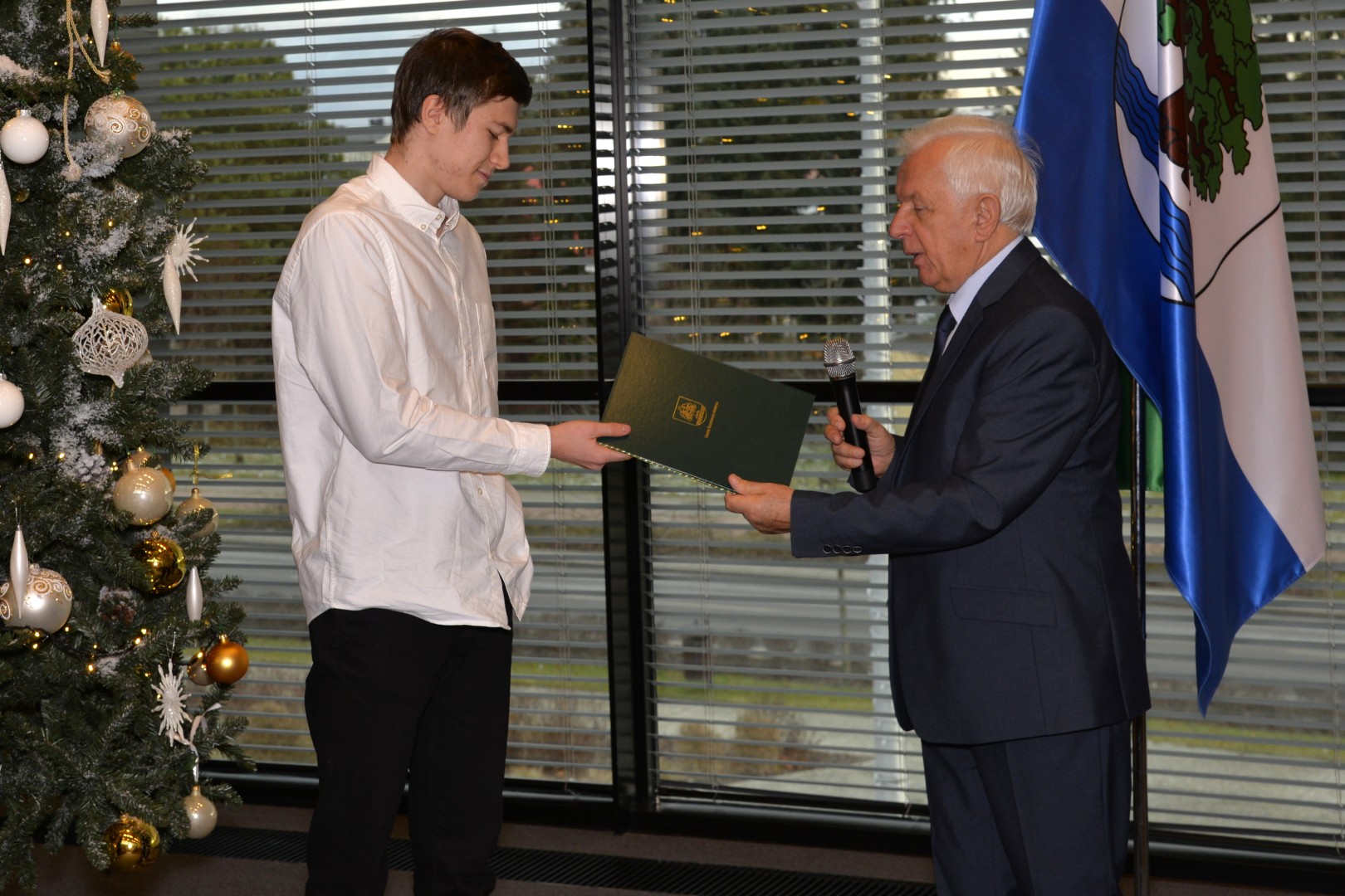 Dwie osoby stoją na tle choinki (nastolatek i mężczyzna). Mężczyzna z prawej (burmistrz) przekazuje młodemu zawodnikowi list gratulacyjny.