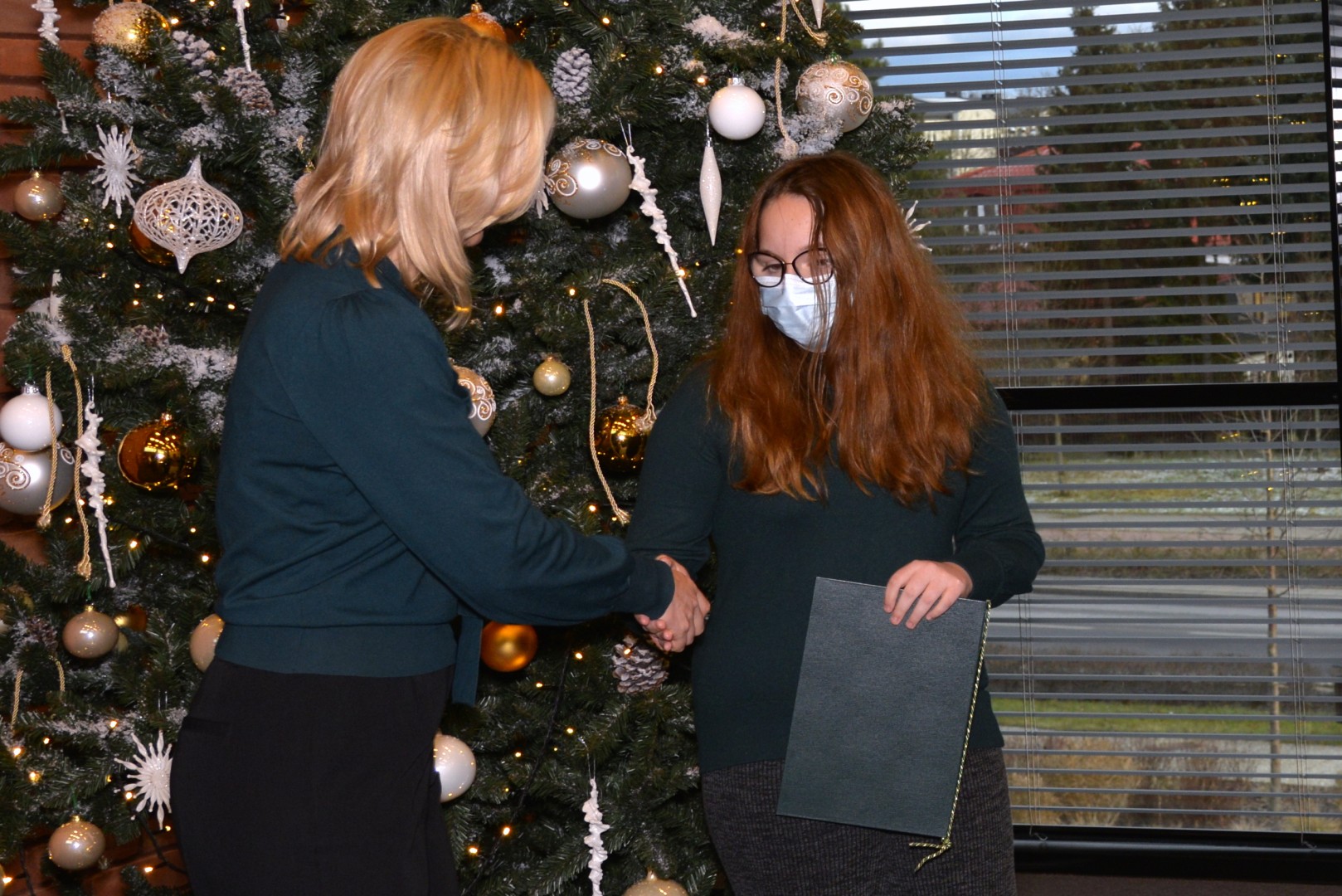 Dwie osoby stoją na tle choinki (dwie kobiety). Kobieta z lewej (przewodnicząca Rady Miejskiej) gratuluje młodej zawodniczce, trzymającej w ręku list gratulacyjny.