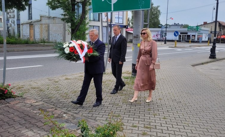 Trzy osoby, przedstawiciele konstancińskiego samorządu, składają kwiaty w miejscu pamięci, osoba w środku trzyma wiązankę kwiatów biało-czerwonych.