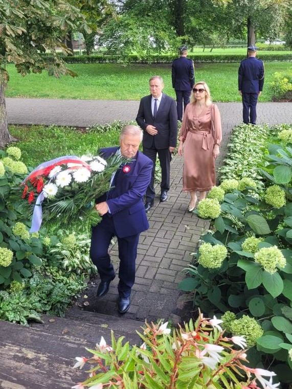 Trzy osoby, przedstawiciele konstancińskiego samorządu, składają kwiaty w miejscu pamięci, osoba w środku trzyma wiązankę kwiatów biało-czerwonych.