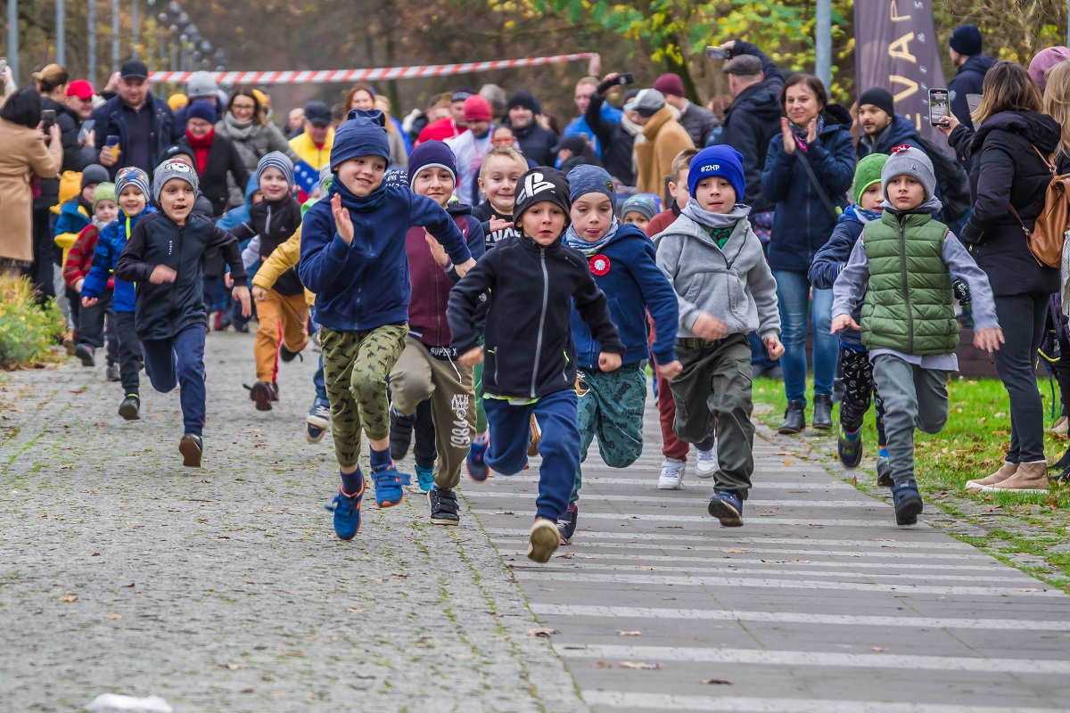 8.	Dzieci biegną chodnikiem w parku, przy chodniku stoją rodzice i robią zdjęcia.