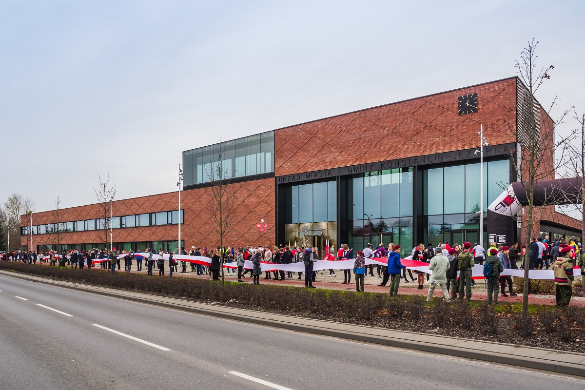 Wzdłuż budynku Urzędu Miasta i Gminy młodzież trzyma 100-metrową biało-czerwoną flagę.