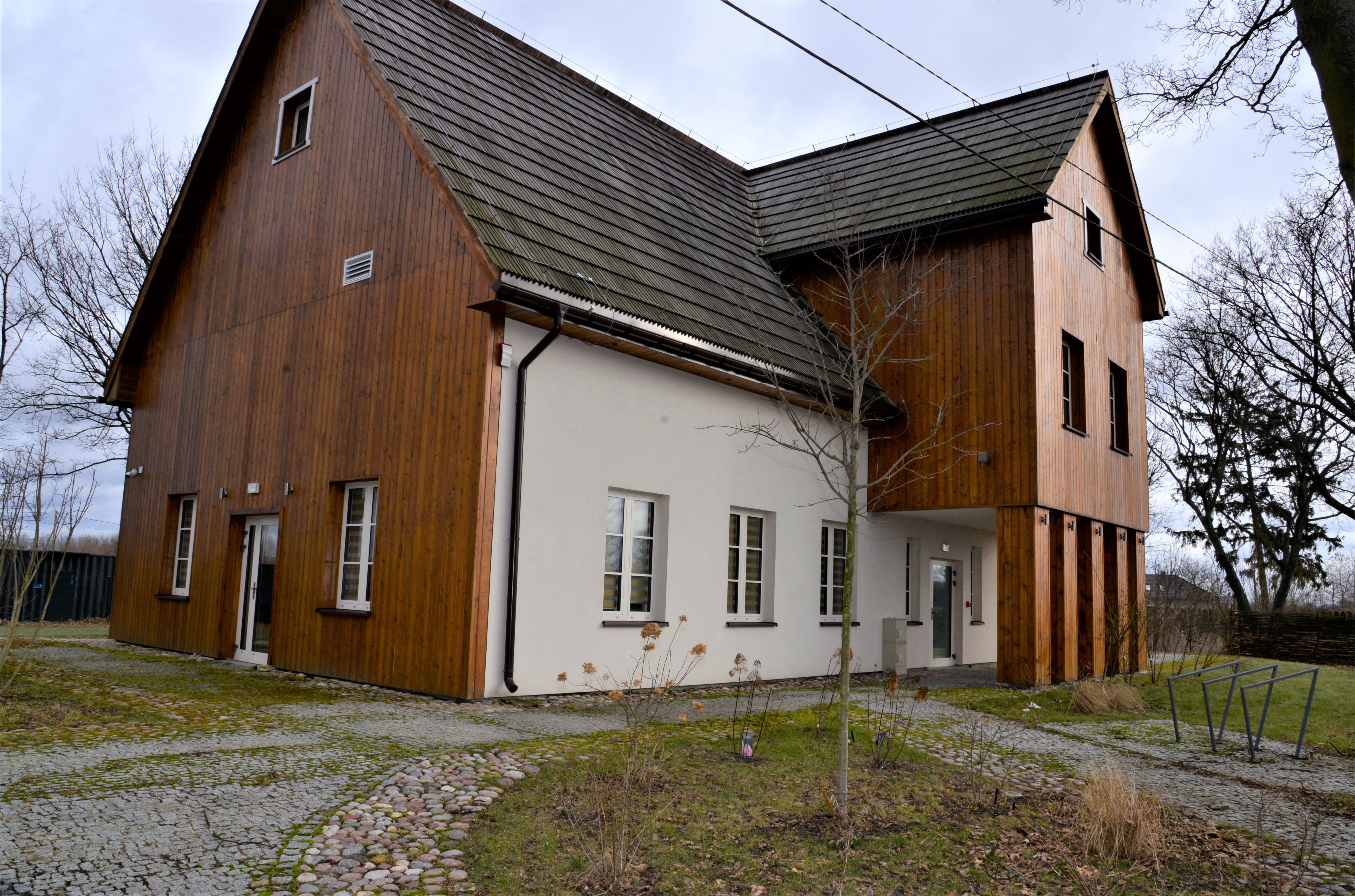 Bydynek z dachem spadzistym widoczny od narożnika, jedna ściana pokryta drewniem, na ścianie po prawej stronie widoczna nadbudówka  pokryta drewnem i podparta kolumnami