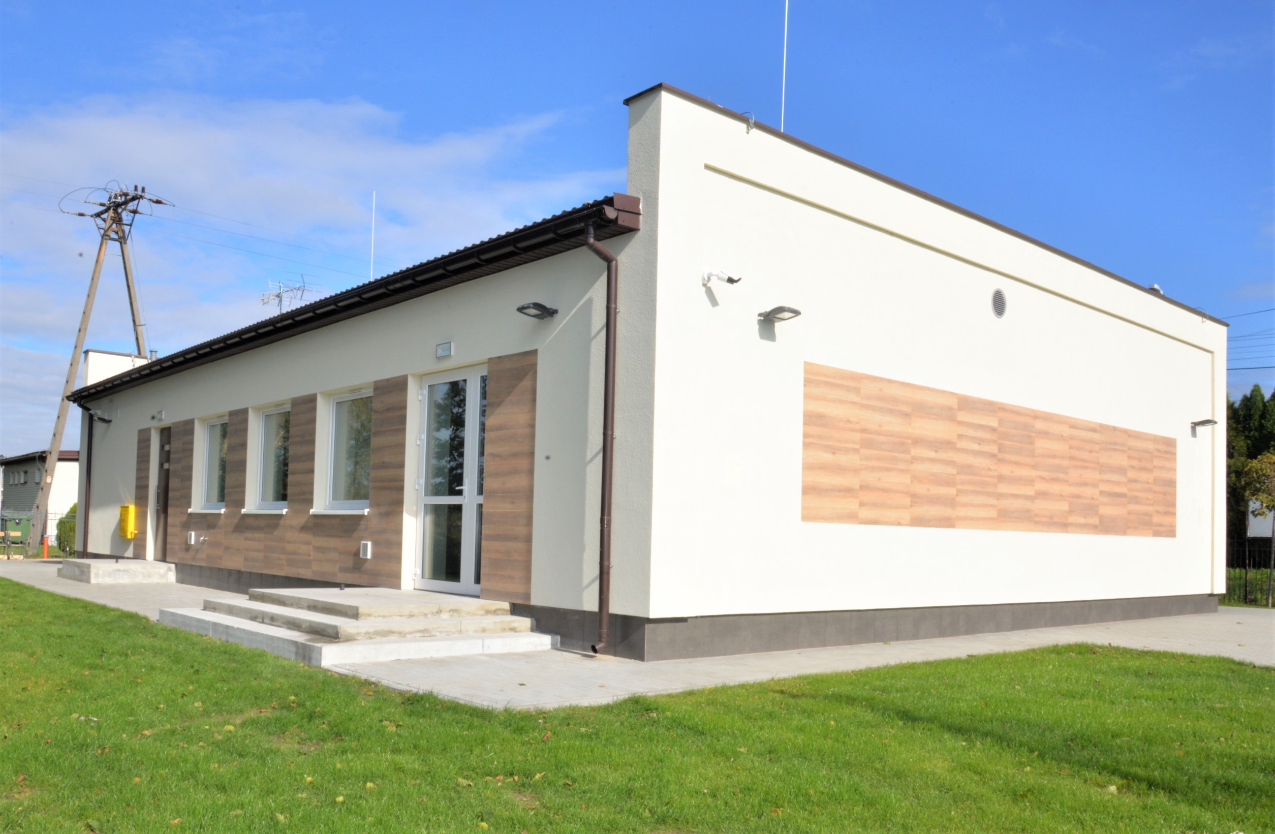 Niski budynek widoczny od narożnika, jasne ściany ze wstawionymi panelami imitacja drewna, w ścianie po lewej okna i drzwi