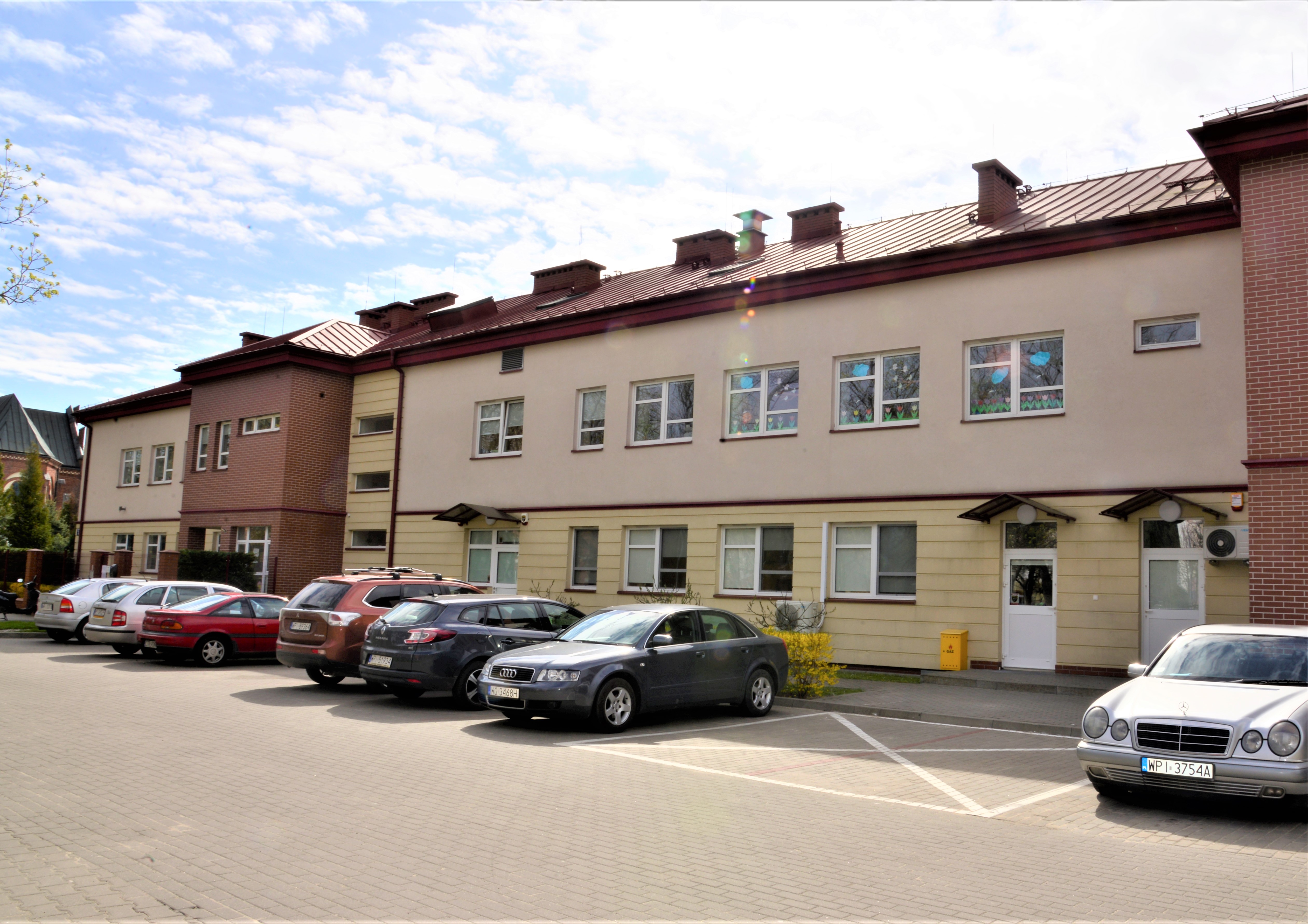 Dlugi, piętrowy budynek, z jasnąelewacją z fragmentami pokrytymi klinkierem, dach ciemny, spadzisty, przed budynkiem stoi kilka samochodów osobowych