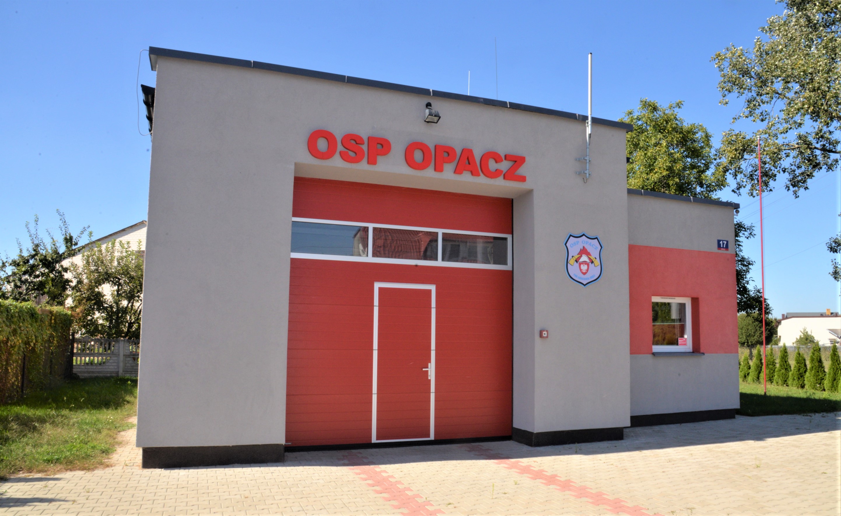 Duży budynek widziany od frontu, pośrodku duża brama wjazdowa w kolorze czerwony, nad nią napis OSP OPACZ w kolorze czerwonym