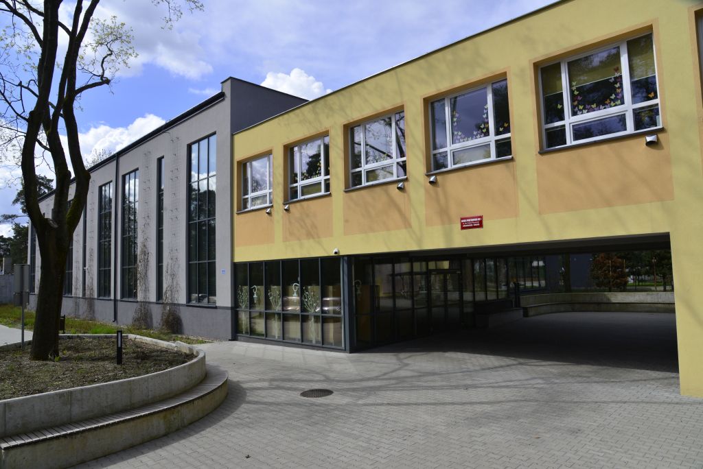 Fragment dużego budynku składającego się z dwóch połaczonych części, część po lewej stronie z dużymi pionowymi oknami, po prawej rodzaj łącznika z wejśćiem na dole