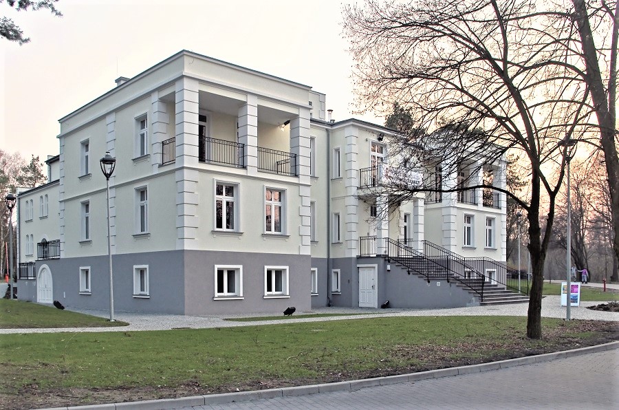 Widok narożny zabytkowego piętrowego budynku z jasną elewacją, na środku frontowej ściany drzwi wejściowe do których prowadzą schody