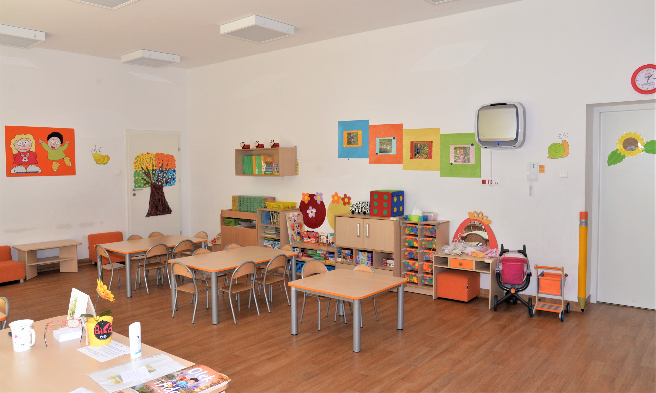 Duża sala z jasnmi ścianami, pośrodku stoją trzy małe stoliki i krzesełeczka dla dzieci, pod ścianą po prawej stroni stoją meble dziecięce wypełnione zabawkami, na ścianach kolorowe i wesołe obrazki