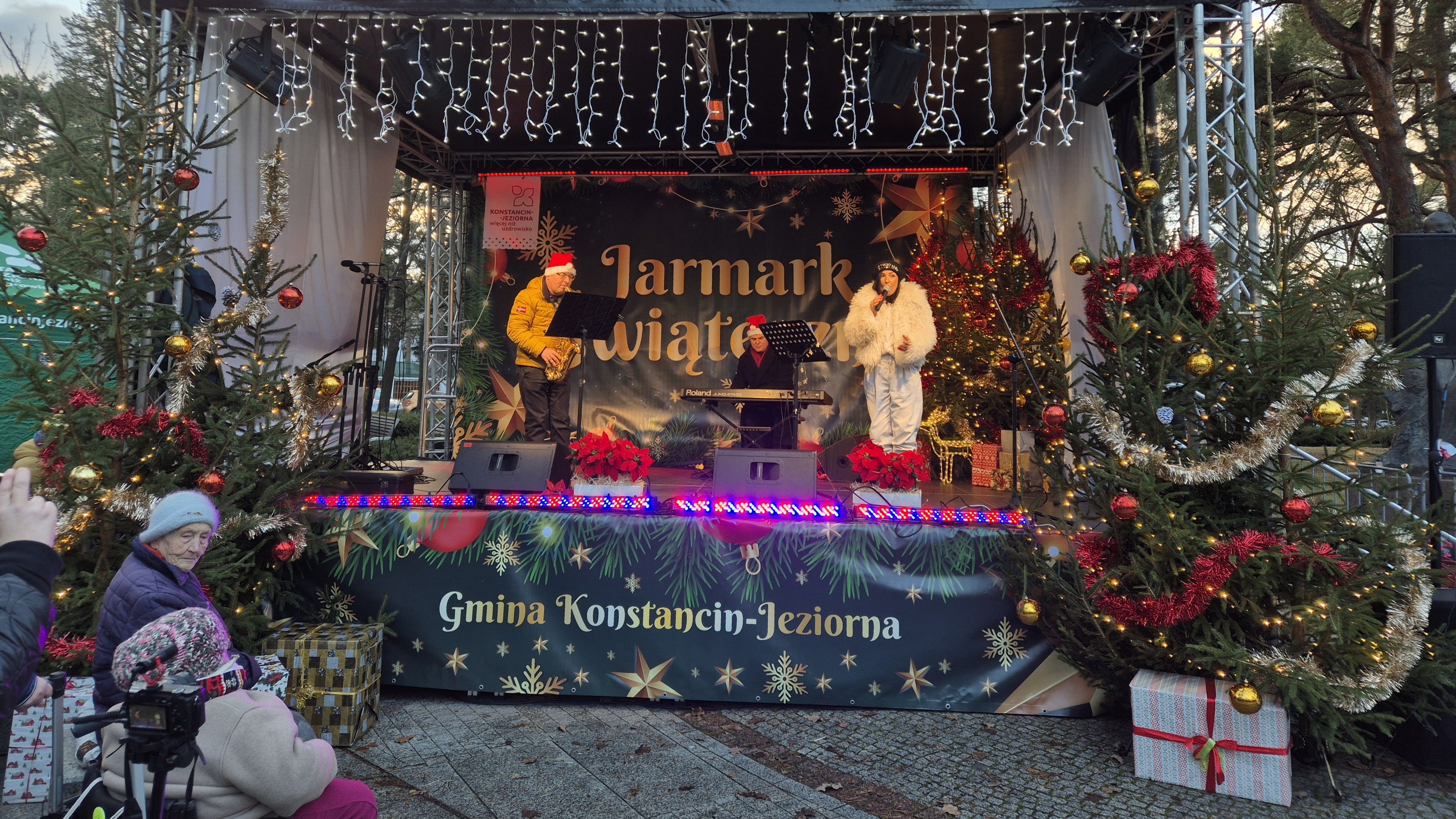 Występ lokalnych zespołów i chórów na scenie, z widocznymi świątecznymi dekoracjami i oświetleniem.