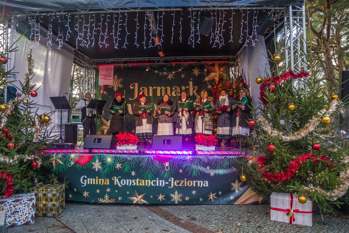 Występ lokalnych zespołów i chórów na scenie, z widocznymi świątecznymi dekoracjami i oświetleniem.