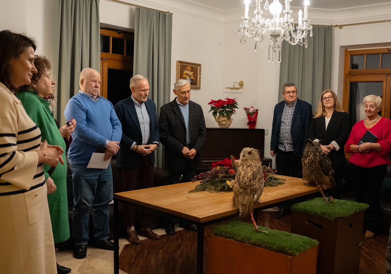 Uczestnicy świątecznego spotkania stoją wokół stołu w sali urzędu, słuchając opowieści o sowach, które znajdują się na środku pomieszczenia.