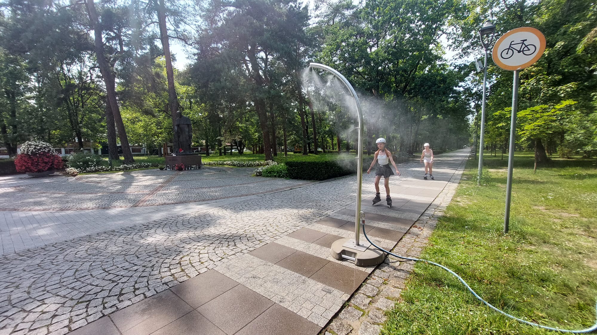 Dzieci na rolkach przejeżdżają przez mgiełkę wodną rozpylaną przez słupek zraszający.