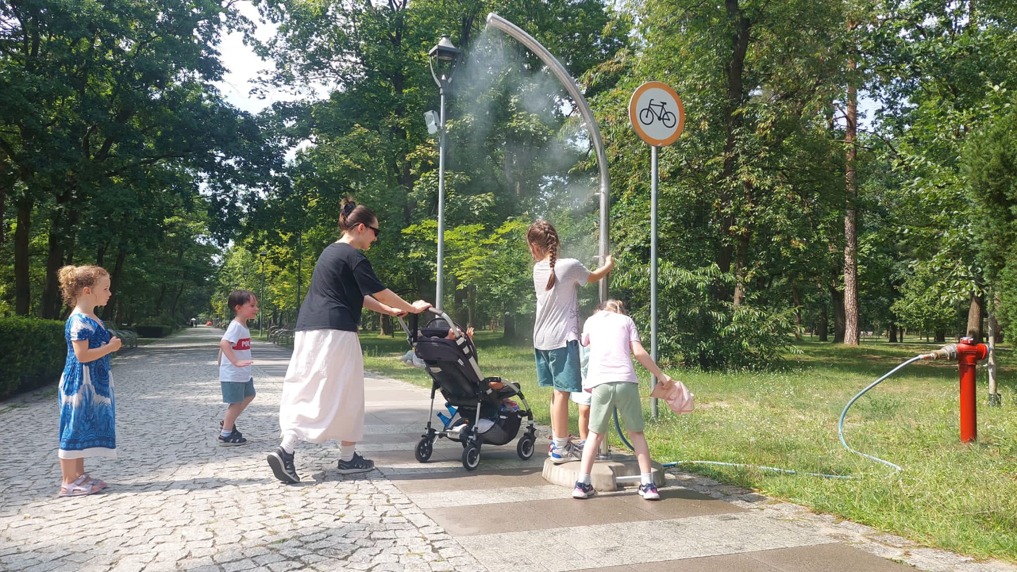 Kobieta z dziećmi korzysta z bramki zraszającej, która rozpylając wodną mgiełkę przynosi ochłodę podczas upalnego dnia.
