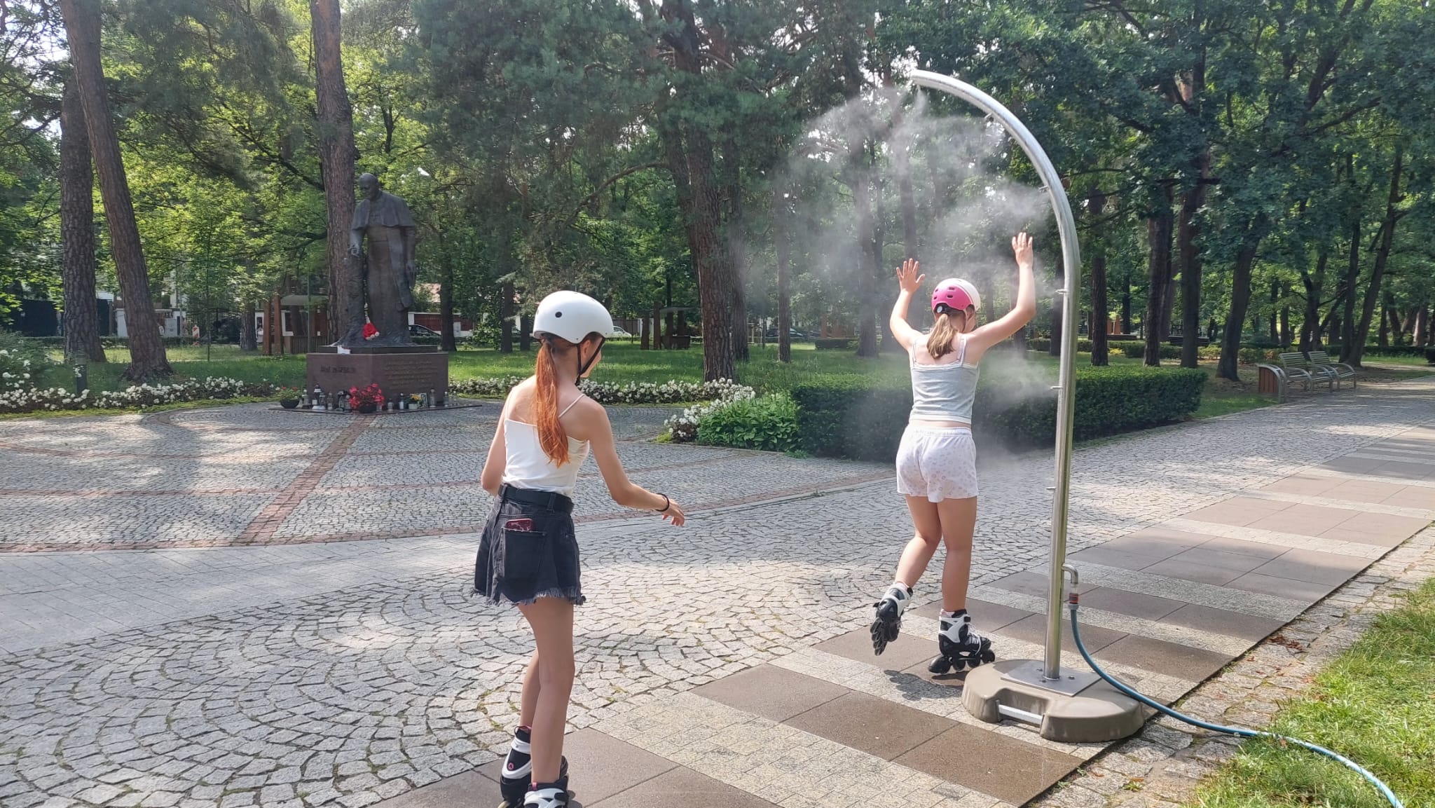 Dwie osoby na rolkach korzystają z ochłody pod słupkiem zraszającym w Parku Zdrojowym.