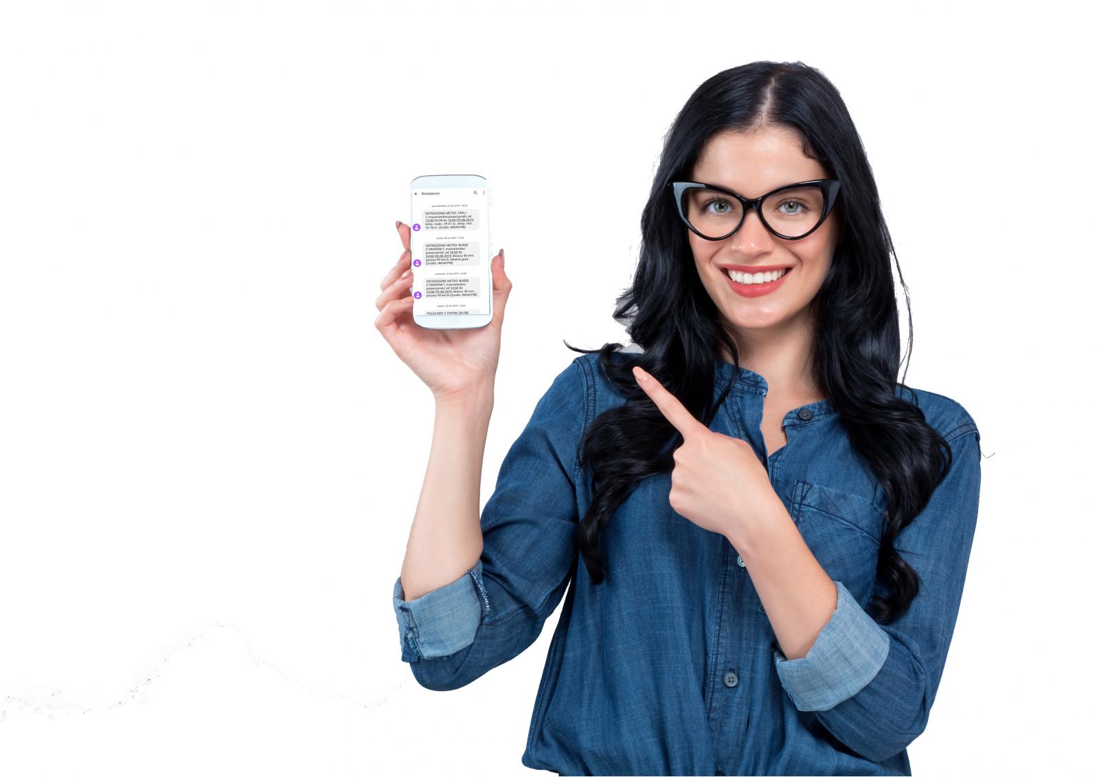 Na zdjęciu młoda uśmiechnięta kobieta (długie czarne włosy, ubrana jest w dżinsową koszulę), która w prawej ręce trzyma telefon komórkowym. Na wyświetlaczu telefonu widać wiadomości od Konstancińskiego Systemu Powiadamiania SMS. 