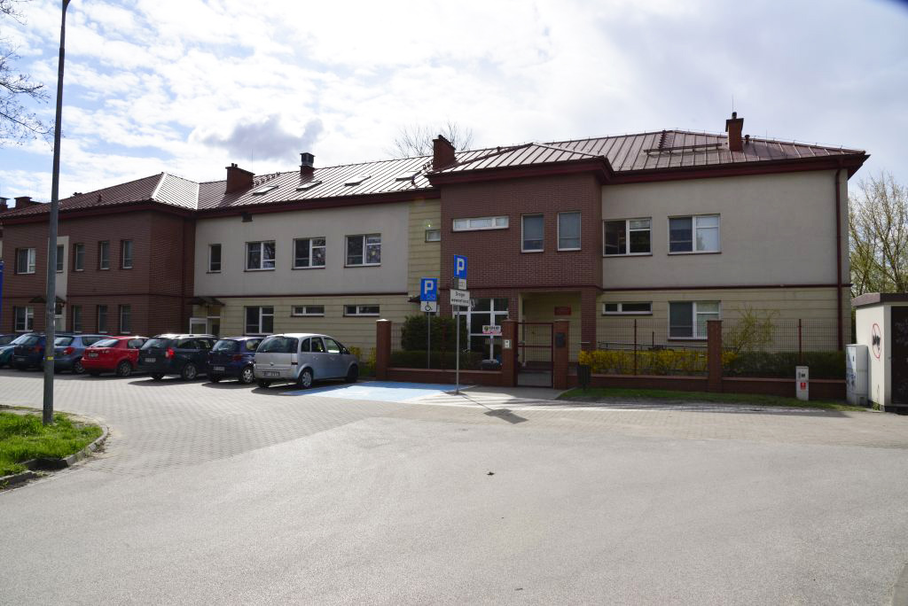 dwukondygnacyjny budynek z jasną elewacja i czerwonymn dachem. Wzdłuż budynku znajduje się rząd zaparkowanych samochodów 