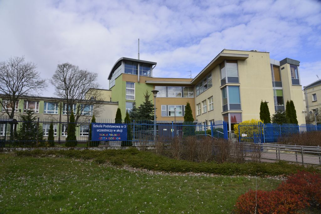 Budynek szkoły z kolorowymi roletami w oknach. Przed szkołą niebieskie ogrodzenie oraz dekoracyjne krzewy.
