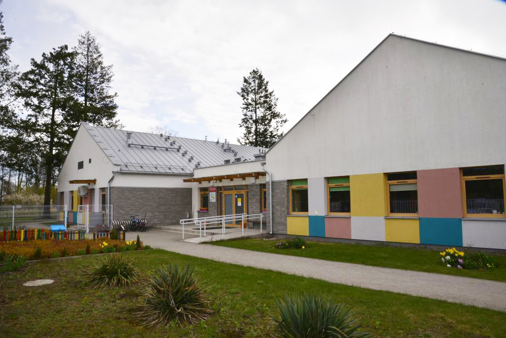 dwa budynki z dwuspadowym dachem połączone łącznikiem, dookoła nasadzenia z roślin. Pomiędzy okanmi znajdują się kolorowe wstawki tynku.