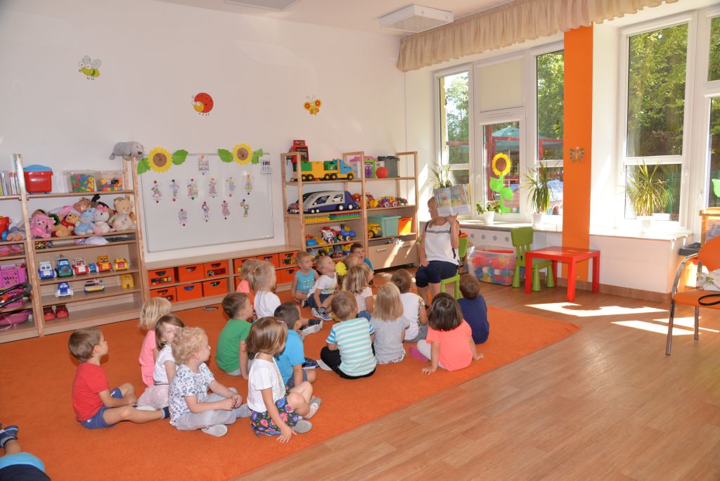 Grupa dzieci siedząca na podłodze słuchająca wychowawcę na sali zabaw przedszkola.