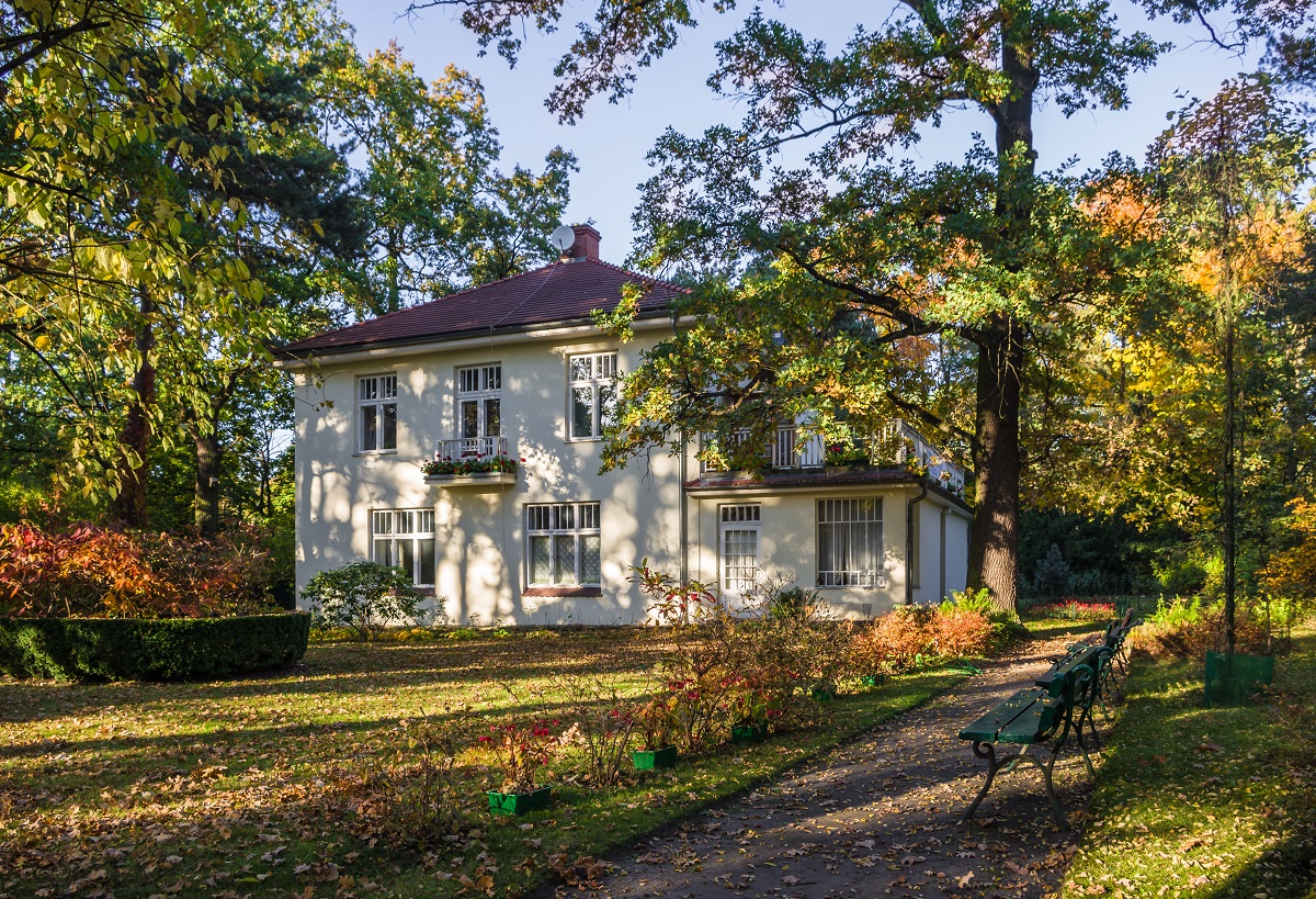 Widok na willę Świt. Biały budynek na planie kwadratu z czerwonym dachem, duża ilością okien. Wokół willi rosną wysokie drzewa. Zdjęcie zrobione w jesienny dzień.