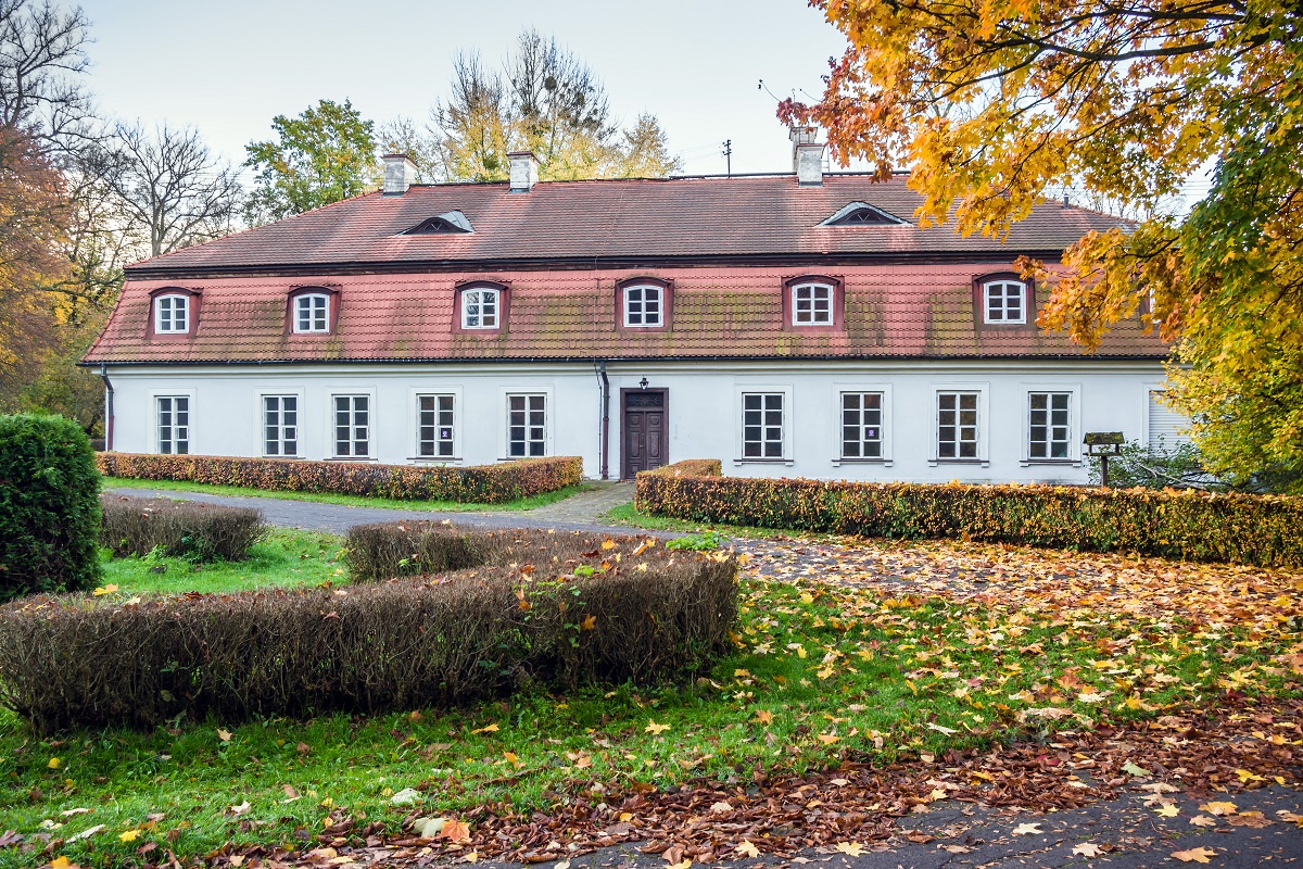 Prostokątny budynek (dwór) z jasną elewacją, dużą ilością okien, z pomarańczowym dwuspadowym dachem. Dokoła rosną wysokie drzewa.  Przed dworem znajdują się trzy naturalne płotki z krzewów. Na trawniku leżą jesienne liście.