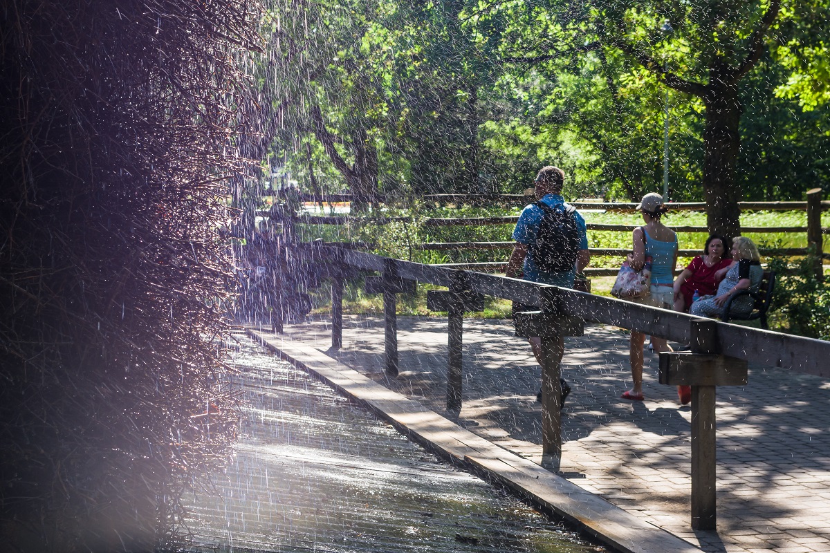 Zdjęcie wykonane w letni dzień i przedstawia drewniany element tężni solankowej, po której skrapla się solankowa woda. Obok tężni spacerują ludzie.