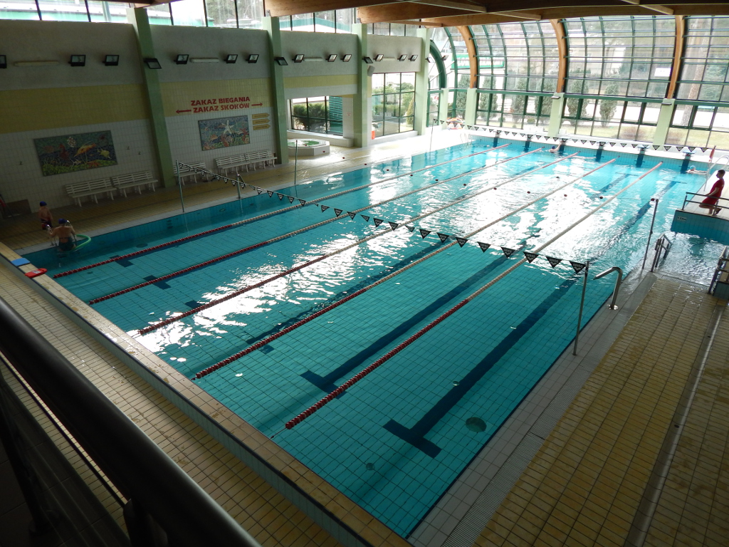 Zdjęcie wnętrza basenu w Centrum Kompleksowej Rehabilitacji (prostokątny basen, podzielony na 6 torów do pływania, na końcu którego znajduje się wysoka szklana ściana, przez którą widać zieleń). 