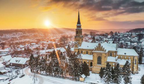 Widok z góry na pokryte śniegiem miasto. W centralnej części, pośród drzew iglastych znajduje się kościół z wysoką wieżą. W tle panorama gór oraz wielobarwne niebo  z zachodzącym słońcem.