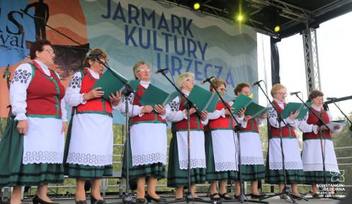 Zdjęcie przedstawia zespół Łurzycanki występujący na scenie podczas Jarmarku Kultury Urzecza. Osiem pań, ubranych jest w tradycyjne stroje z regionu. W rękach trzymają śpiewniki. 