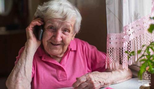 Starsza kobieta siedzi przy stole, lewa dłoń oparta na blacie, prawą dłonią trzyma przy uchu aparat telefoniczny