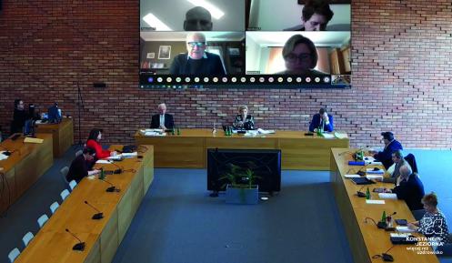 Sala obrad rady miejskiej, widok z góry, na środku ekran z wyświetlanymi ekranami komputerowymi z radnymi, pod nimi duży stół z osobami, po prawej i lewej przy długich stołąch siedzą osoby