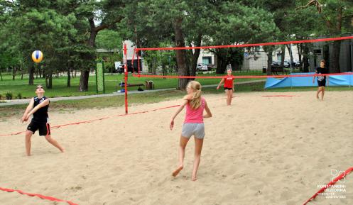 Boisko do gry w piłkę plażową z wyznaczonymi liniami do gry oraz założoną siatką. Dwie pary starszych dzieci gra w siatkówkę. Wokół rosną wysokie drzewa. 